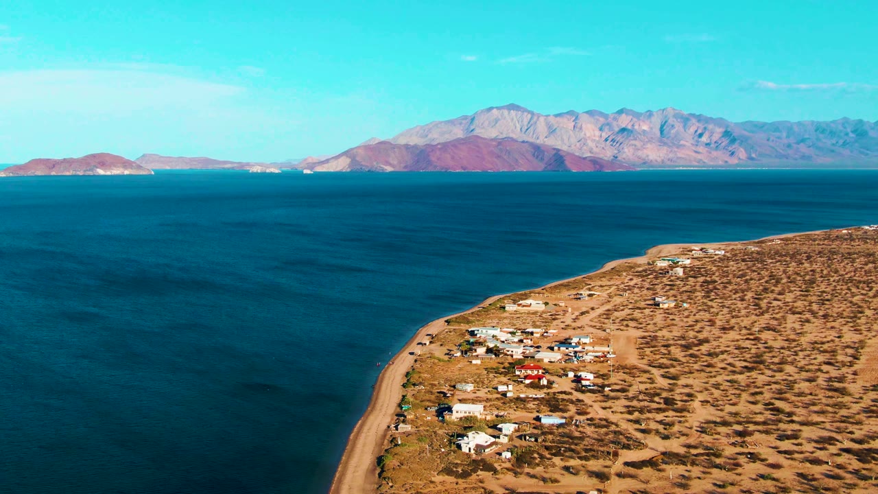 Aerial frame over Bahia De Los Angeles with more of the settlement visible at lower right, a narrow sandy beach along the water, a broad blue bay, distant mountains, and no visible people.