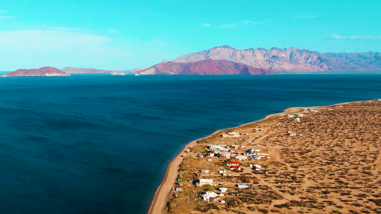 Wide aerial coastline with small buildings, open ocean, and mountain horizon.