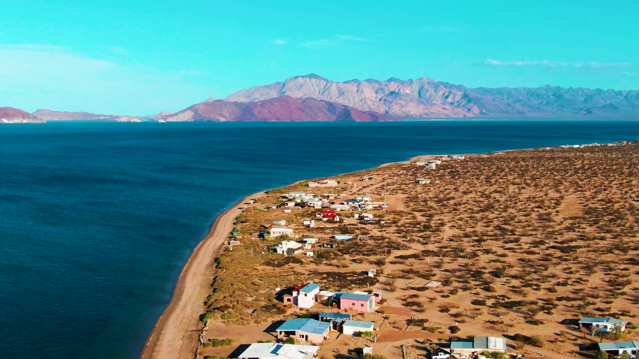 Opening aerial frame over Bahia De Los Angeles, Mexico with blue ocean, a curved sandy shoreline, scattered coastal homes on dry terrain, distant mountains, and no visible people.