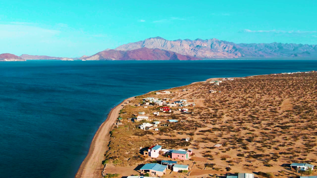Early aerial view of Bahia De Los Angeles with the shoreline running diagonally past small houses, blue ocean filling the left side, distant mountains, and no visible people.