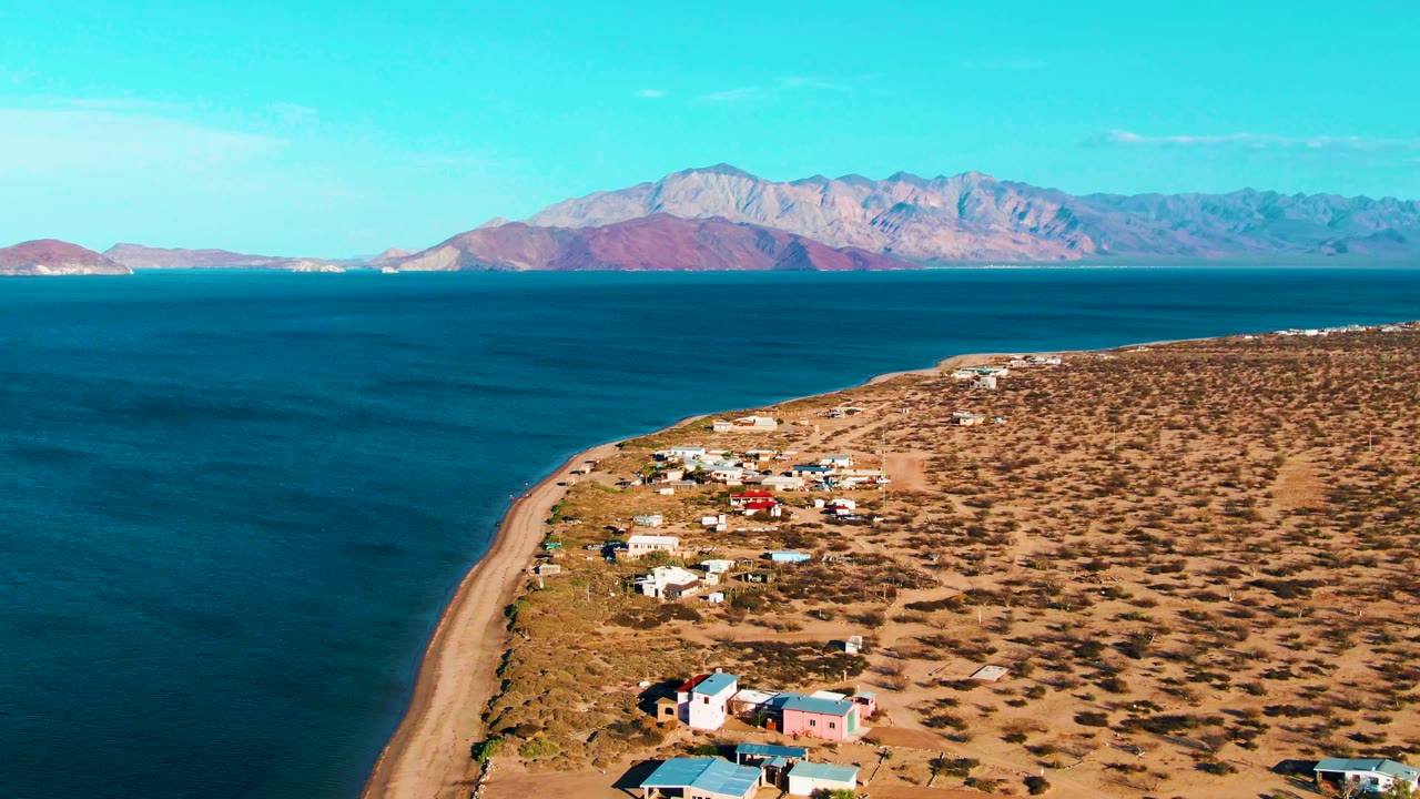 Aerial frame over Bahia De Los Angeles showing deeper blue water on the left, a narrow sandy beach, homes spread across dry brush, mountains across the bay, and no visible people.