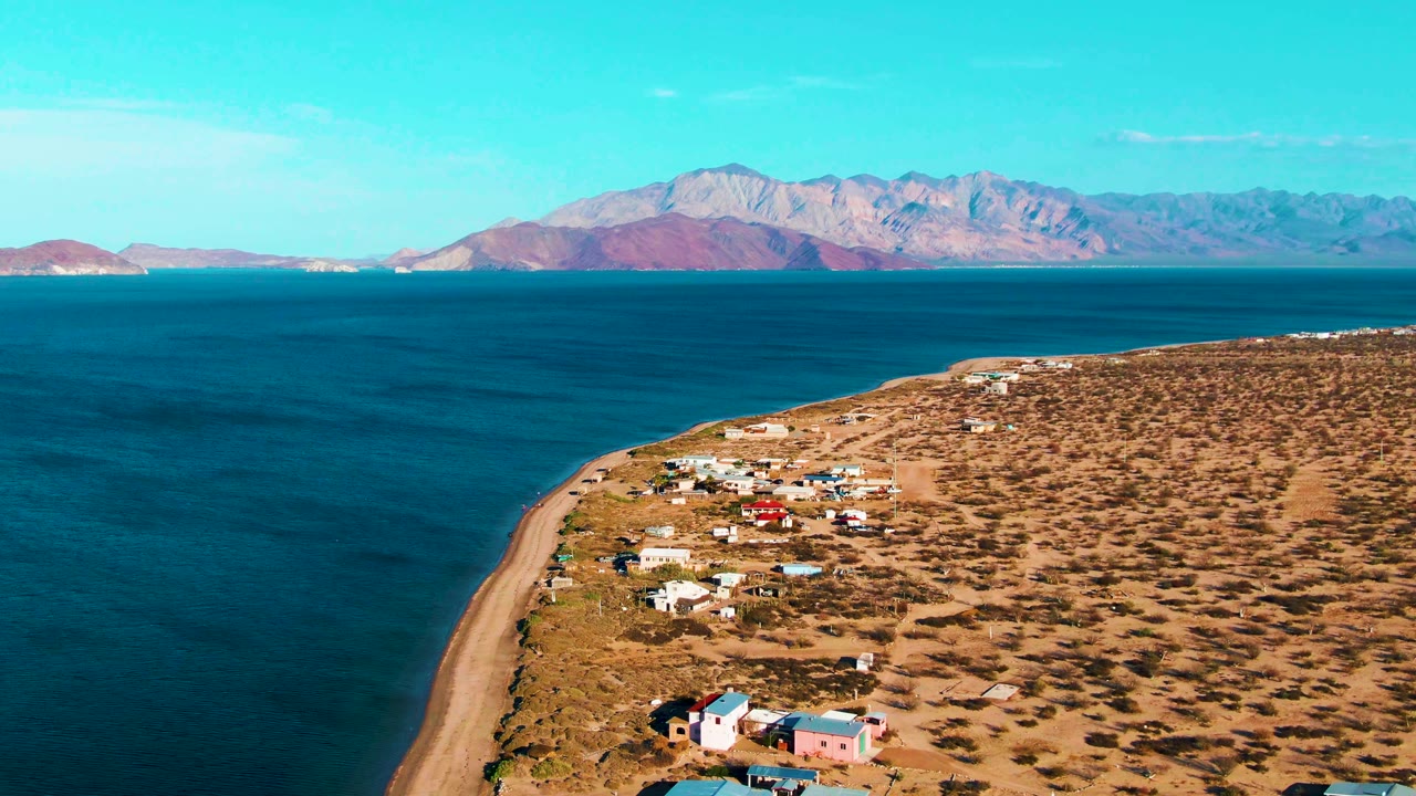 Wide aerial coastline with scattered buildings and distant mountains under blue sky.