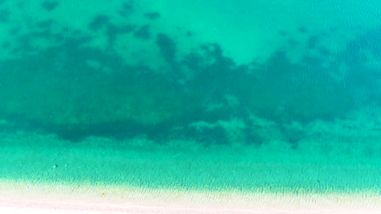 Aerial-looking frame of turquoise sea over rippled sand in Bahia De Los Angeles, Mexico, with irregular darker underwater shapes near the upper edge and no visible people.