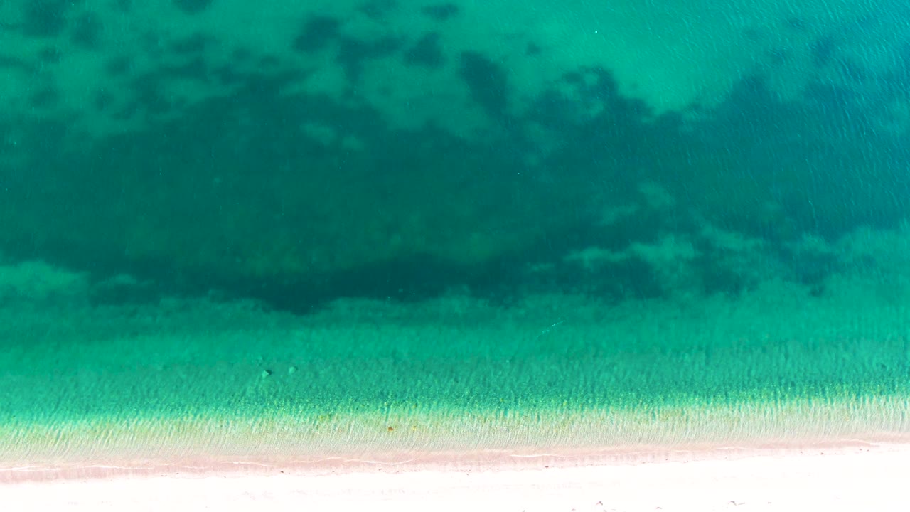 Overhead view of turquoise ocean water and a bright sandy beach in Bahia De Los Angeles, with dark underwater patches offshore and no visible people.