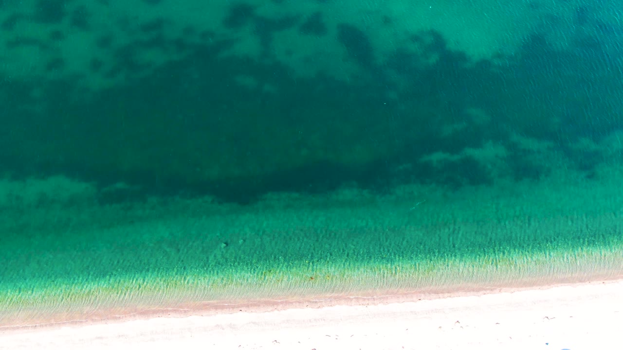 Aerial shoreline frame from Bahia De Los Angeles showing turquoise shallows, pale sand, and broad dark underwater patches, with no visible people.