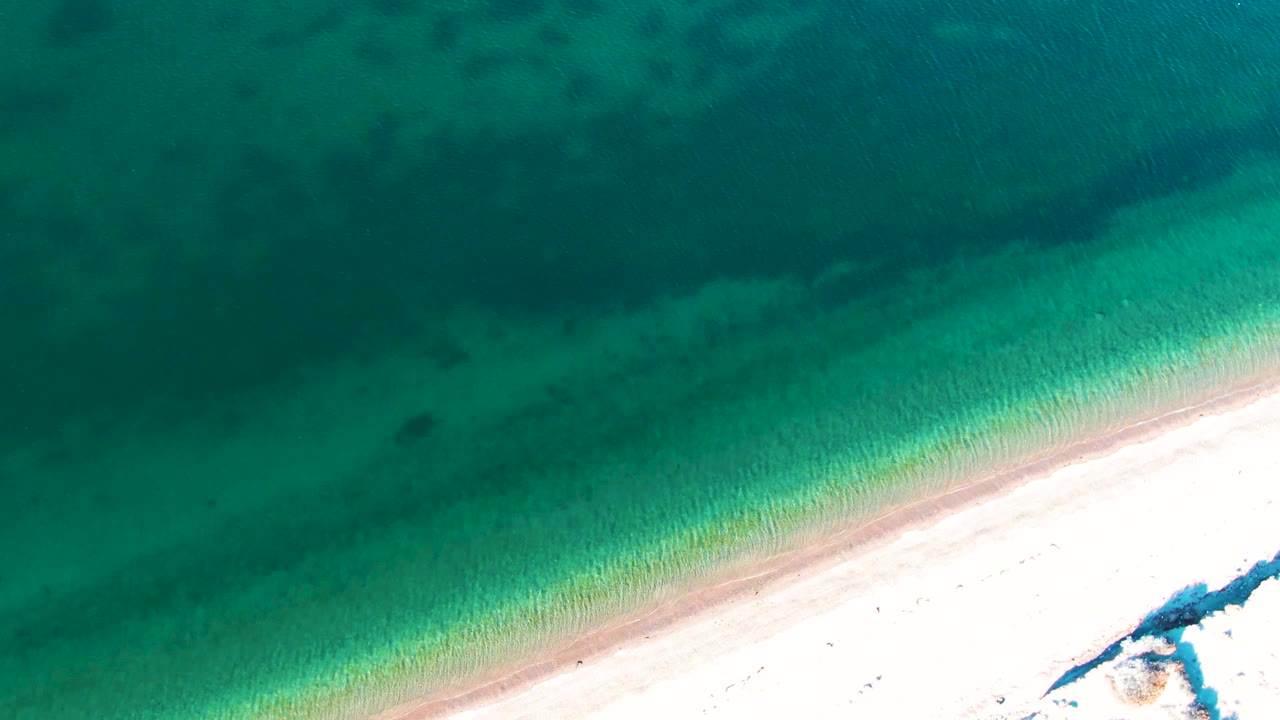 Overhead view of turquoise water crossing a pale sandy shoreline in Bahia De Los Angeles, with darker submerged patches offshore and no visible people.