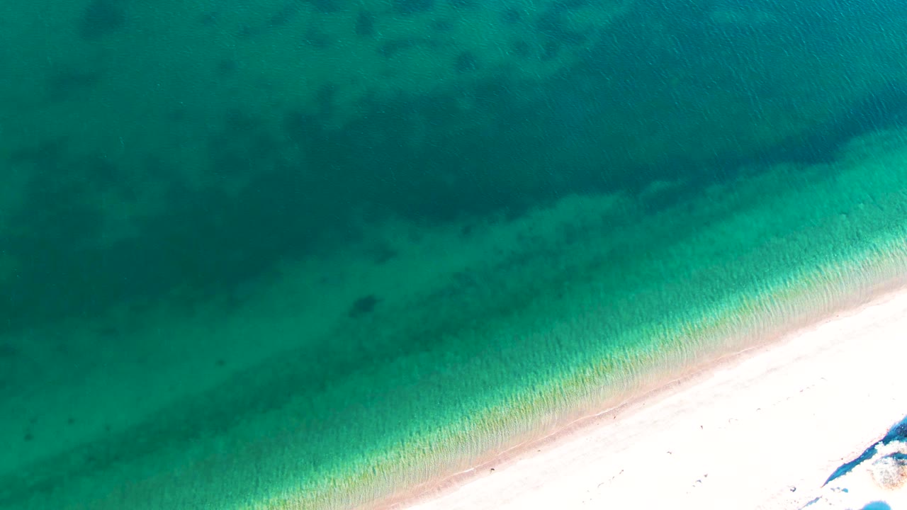 Aerial view of shallow turquoise water and pale sand in Bahia De Los Angeles, with a thin foamy edge near the lower right and no visible people.