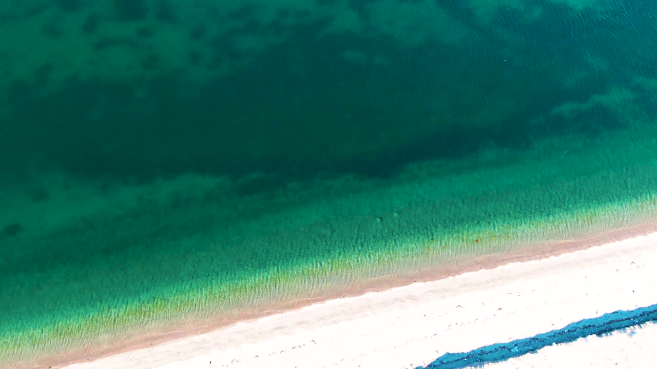 Opening overhead frame of Bahia De Los Angeles turquoise water grading into pale sand, with a diagonal shoreline and no visible people.