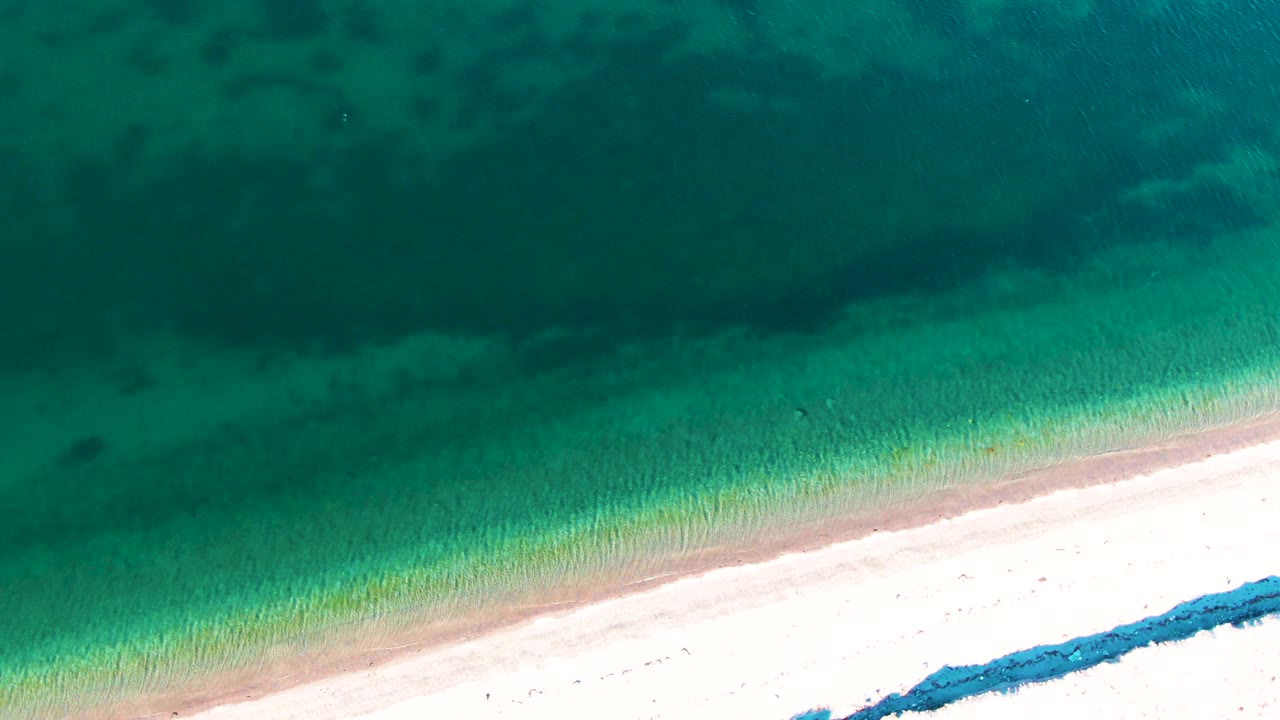 Second overhead frame in Bahia De Los Angeles showing shallow green water, a narrow wet sand band, bright beach, and no visible people.
