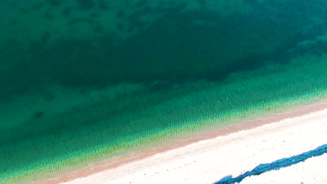 Third frame of the Bahia De Los Angeles beach from above, with the surf line shifted slightly across the diagonal sand edge and no visible people.