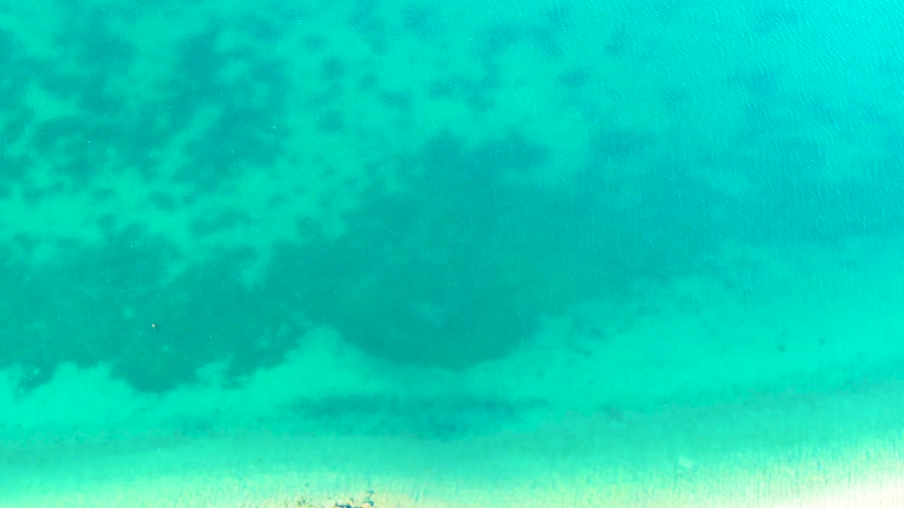 A top-down Bahia De Los Angeles frame shows blue-green water, a pale sandy band near the lower edge, soft white surface wash, and no visible people.