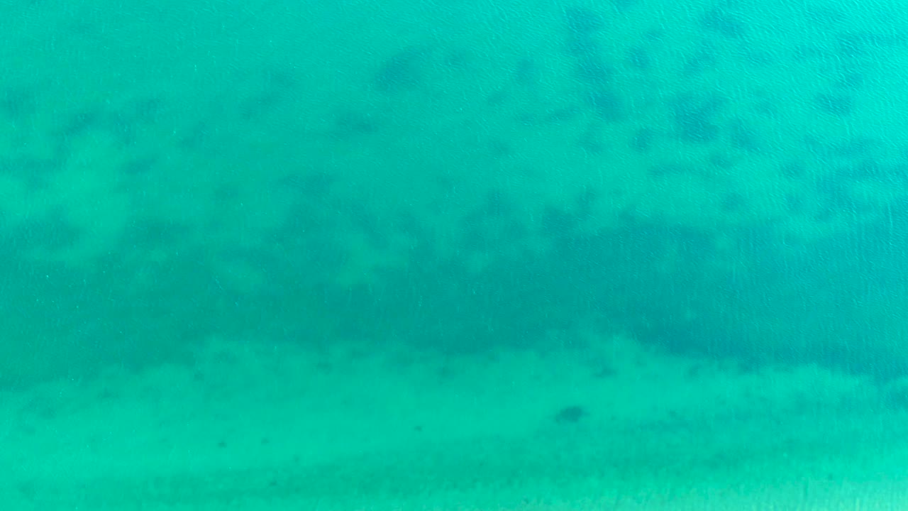 At 0:01, the Bahia De Los Angeles underwater view remains filled with turquoise water, pale sand, and small dark fish marks spread across the frame with no visible people.