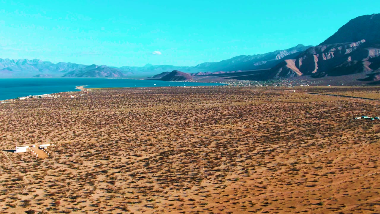 At 0:01, the Bahia De Los Angeles aerial view holds on a sparse brown plain with a small pale structure low in frame, blue water at left, dark mountains at right, and no visible people.
