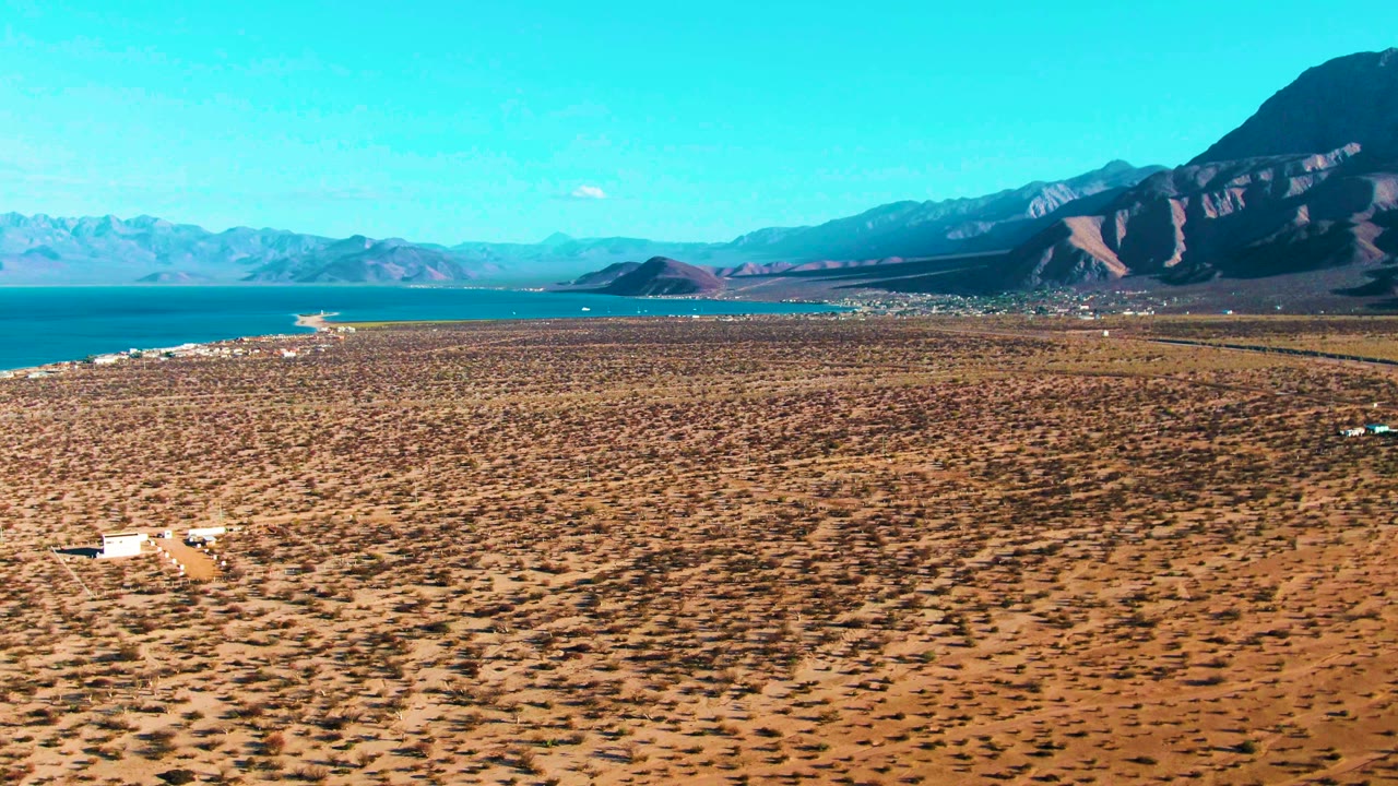 At 0:03, the Bahia De Los Angeles aerial composition keeps the brown arid plain dominant, with blue water along the left edge, mountain ridges beyond, and no visible people.