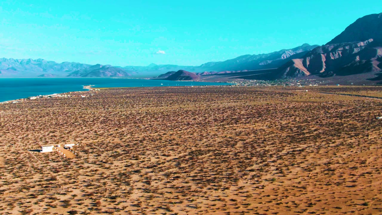 A high aerial view in Bahia De Los Angeles shows a white vehicle low in frame on a dry scrub plain, with blue water to the left, mountains on the horizon, and no visible people.
