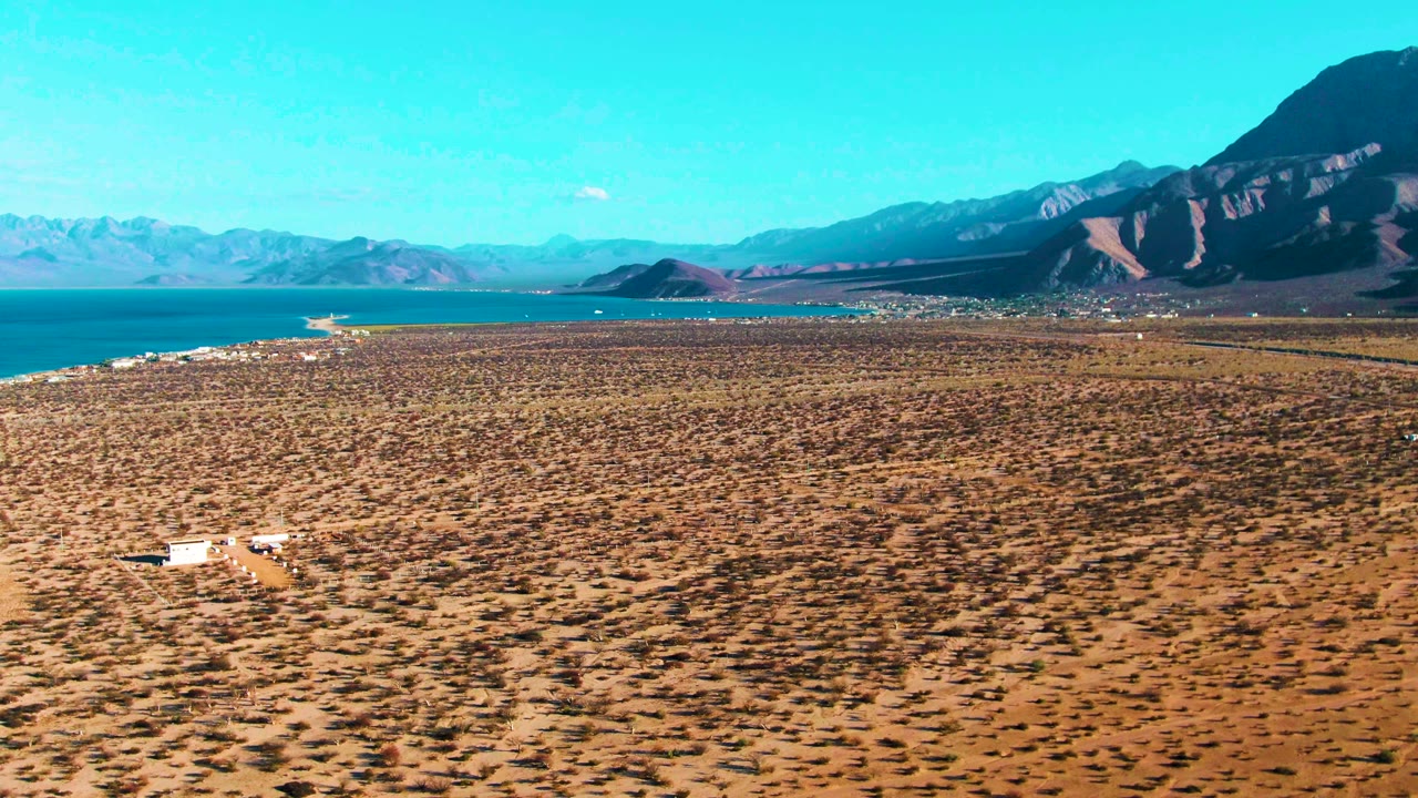 The white vehicle appears slightly farther right on the Bahia De Los Angeles plain, surrounded by sparse brush, with the bay, distant mountains, and clear sky behind it and no visible people.