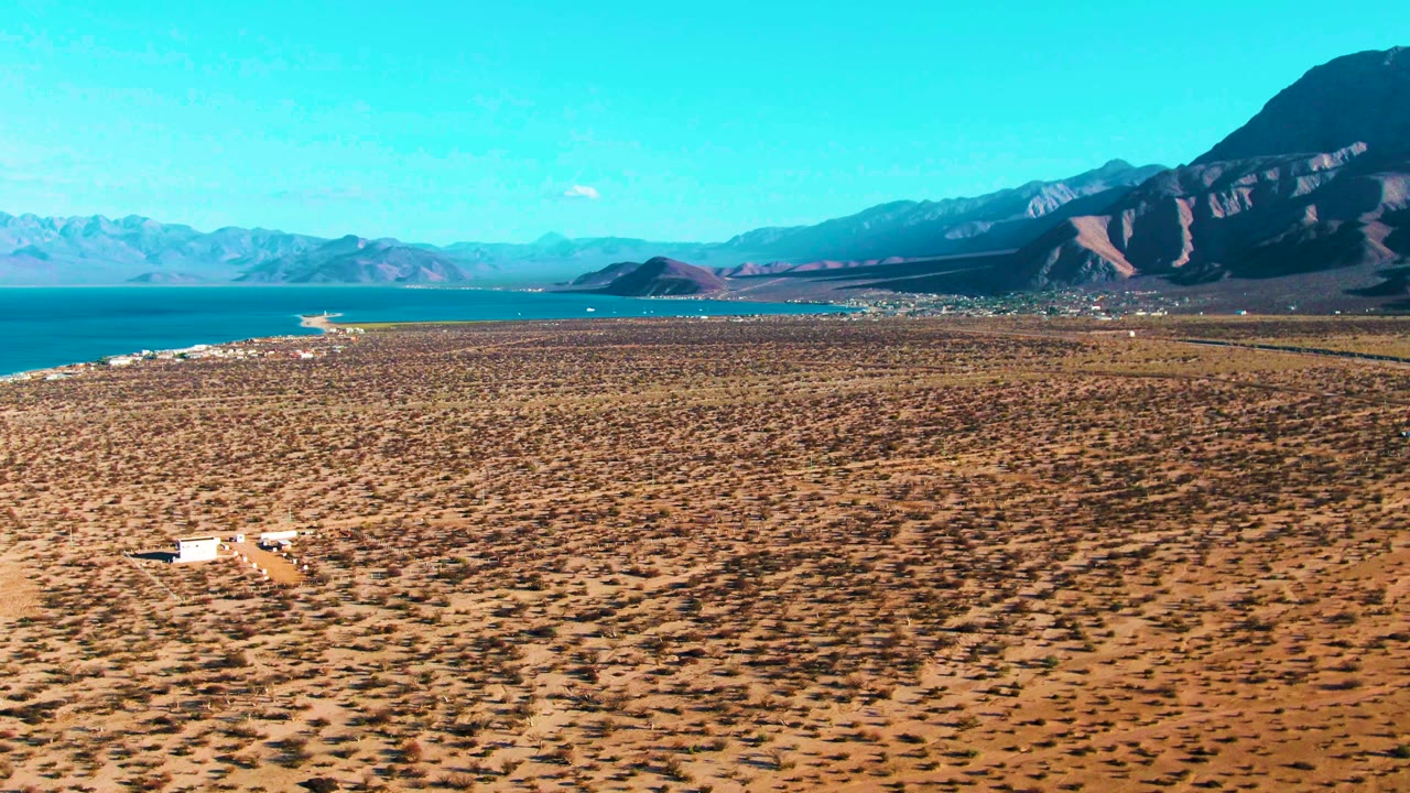 Aerial footage over Bahia De Los Angeles frames the vehicle near the lower left-to-center area, with patterned scrubland leading to blue water, shoreline buildings or light structures in the distance, mountains, and no visible people.