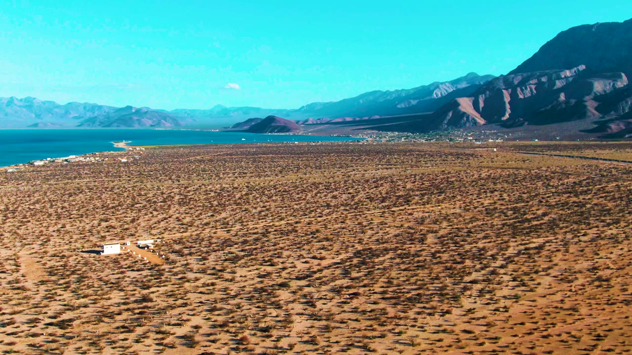 Wide aerial view in Bahia De Los Angeles, Mexico showing a small white vehicle near the lower left, dry desert scrub, blue water, distant mountains, and no visible people.