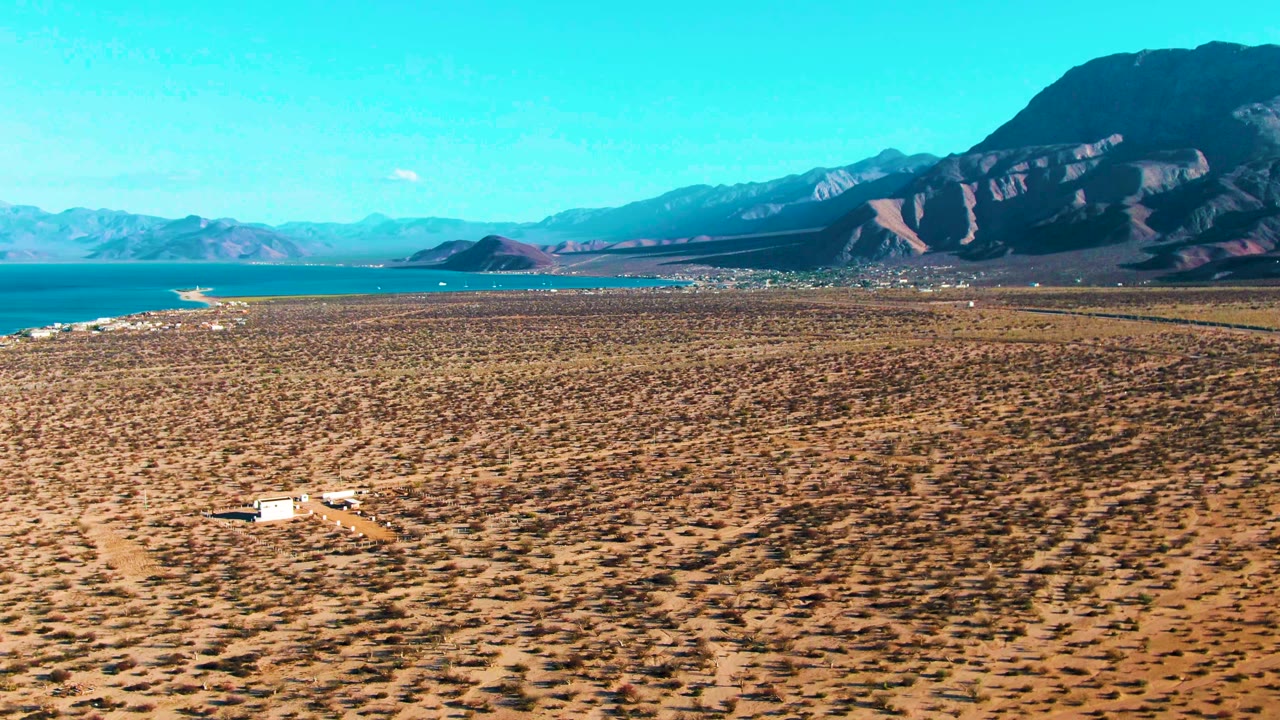 At 0:00 in Bahia De Los Angeles, a white vehicle sits low in frame on a dry scrub plain with blue water at left, dark mountains beyond, and no visible people.
