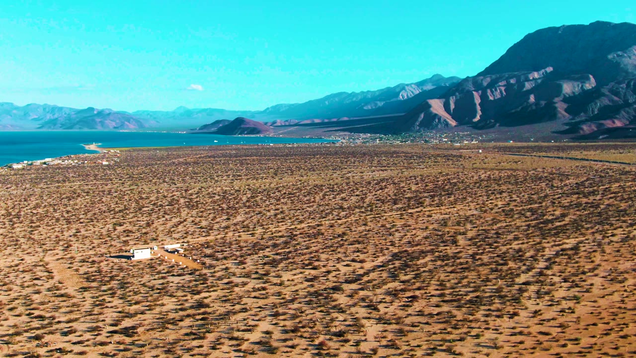 At 0:01 in Bahia De Los Angeles, the white vehicle appears slightly right of its first position on the scrub-covered plain, with the coastline and mountain range still visible and no visible people.