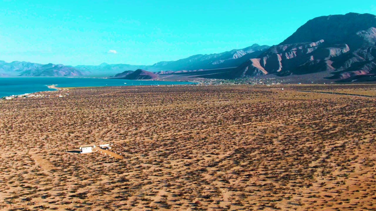 At 0:02 in Bahia De Los Angeles, an elevated view holds the white vehicle near the lower frame, a faint dirt road across the brown plain, blue sea at left, mountains behind, and no visible people.