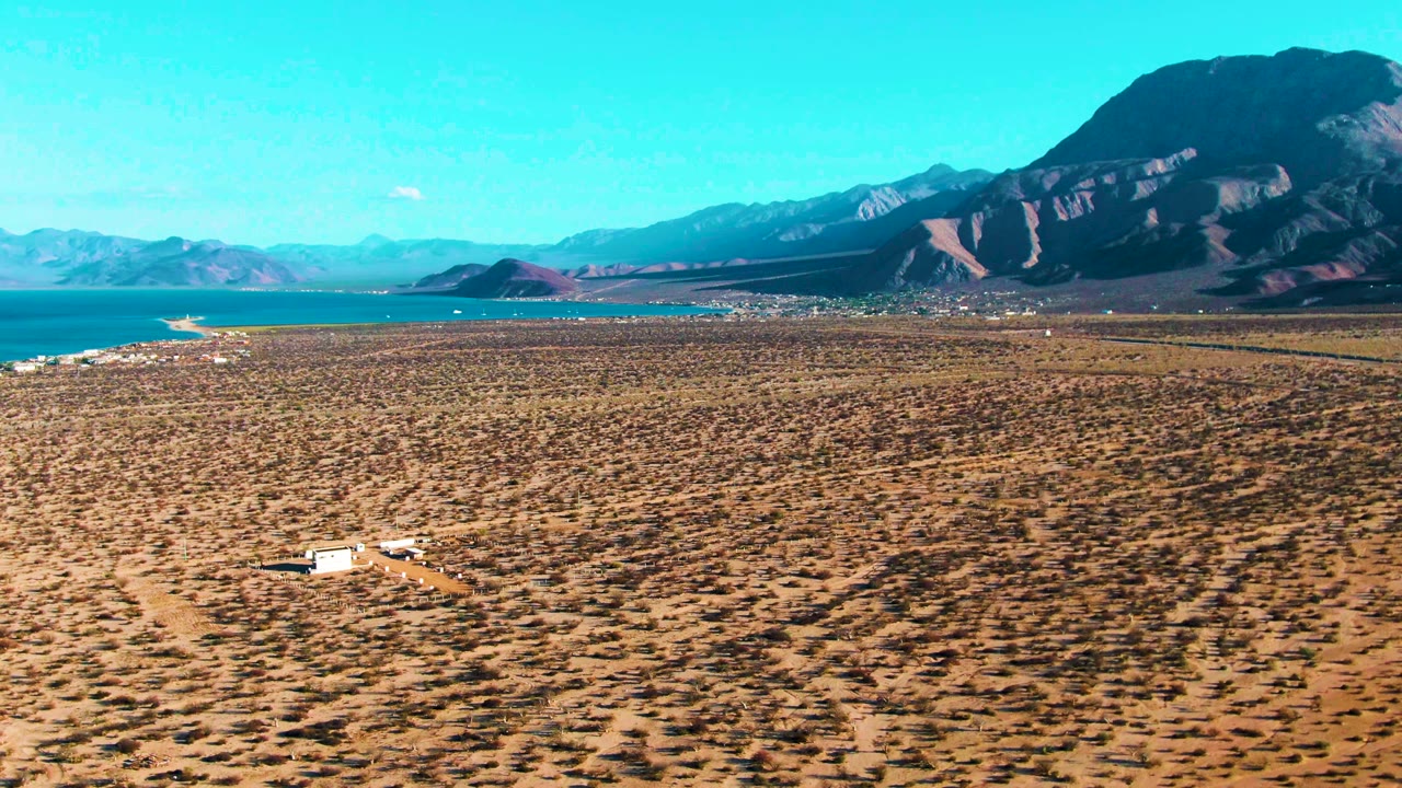 At 0:03 in Bahia De Los Angeles, the white vehicle remains in the lower left area of the dry plain while turquoise water, a pale shoreline, and layered mountains fill the distance, with no visible people.
