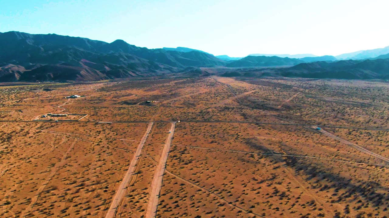 Aerial view over Bahia De Los Angeles desert terrain with dirt roads crossing the foreground, low scrub, distant mountains, bright sky, and no visible people.