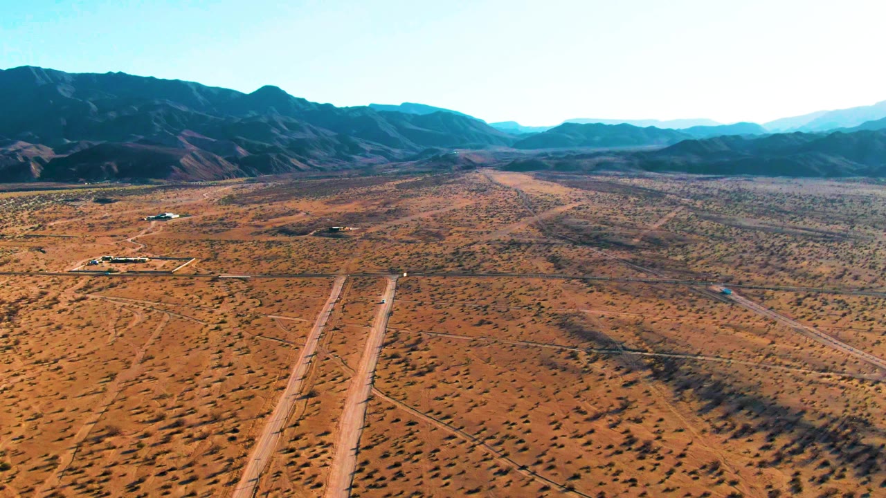 High aerial frame in Bahia De Los Angeles showing a long dirt track through dry brown terrain, scattered shrubs, distant mountain ridges, and no visible people.
