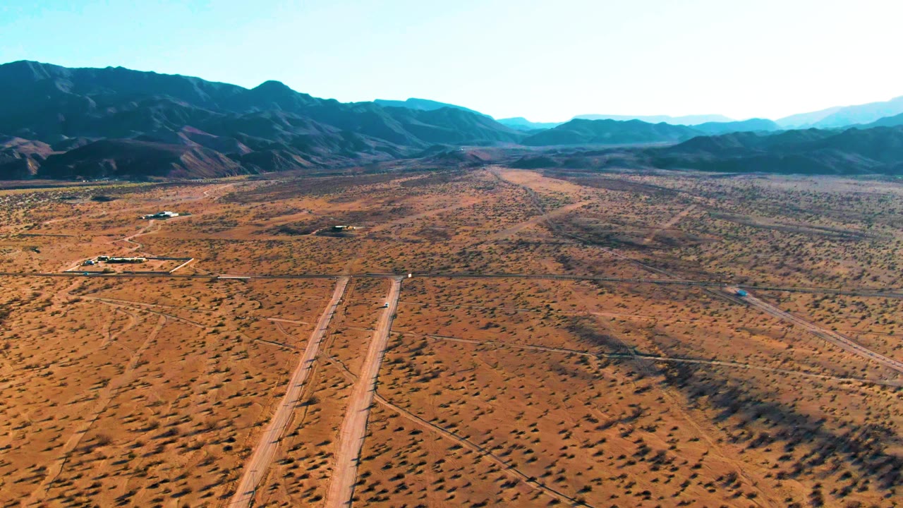 Wide aerial view of Bahia De Los Angeles desert with a pale dirt road in the foreground, a small dark vehicle shape, broad scrubland, dark mountains, and no visible people.