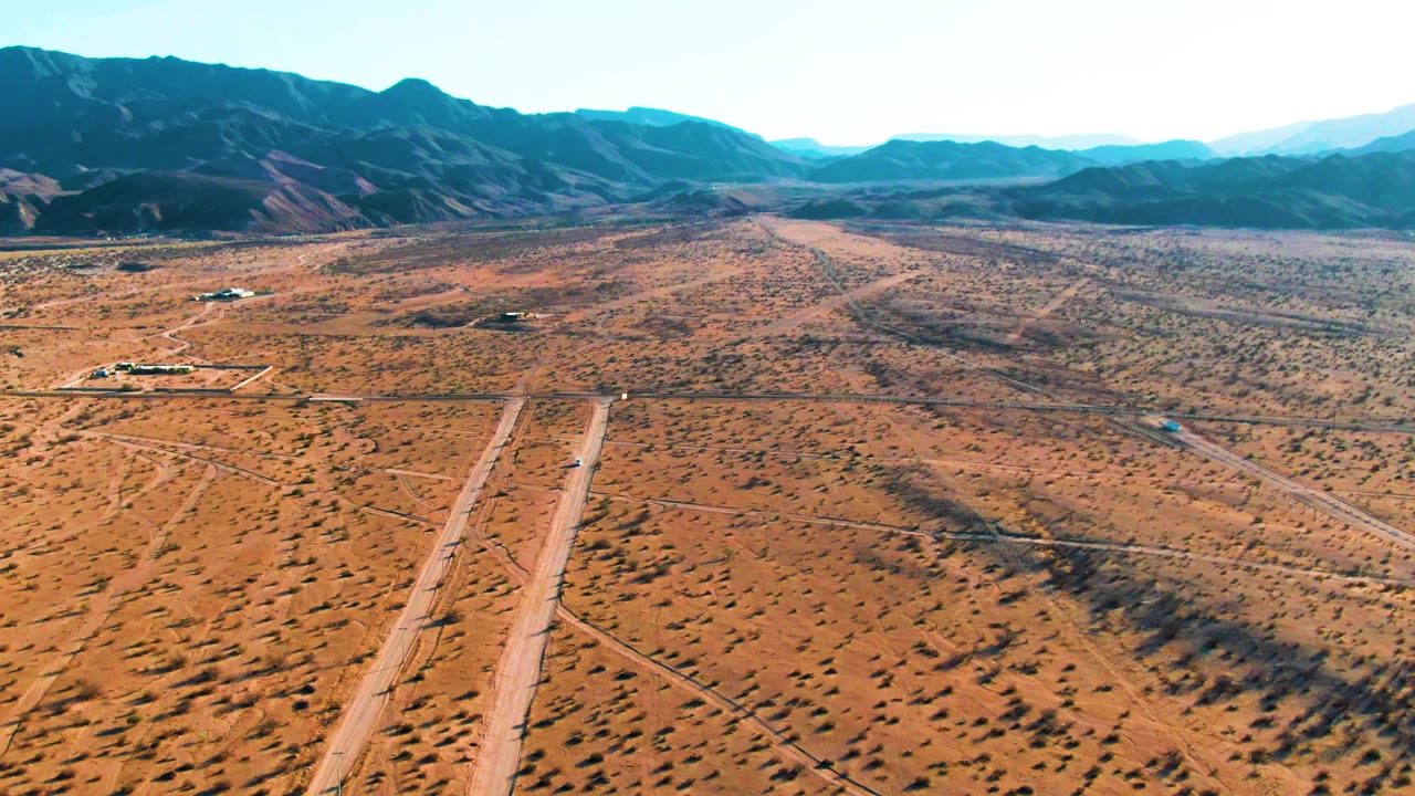 At 0:00, a small aircraft sits low on a desert airstrip in Bahia De Los Angeles, with sandy scrubland, dirt tracks, distant blue mountains, bright sky, and no visible people.