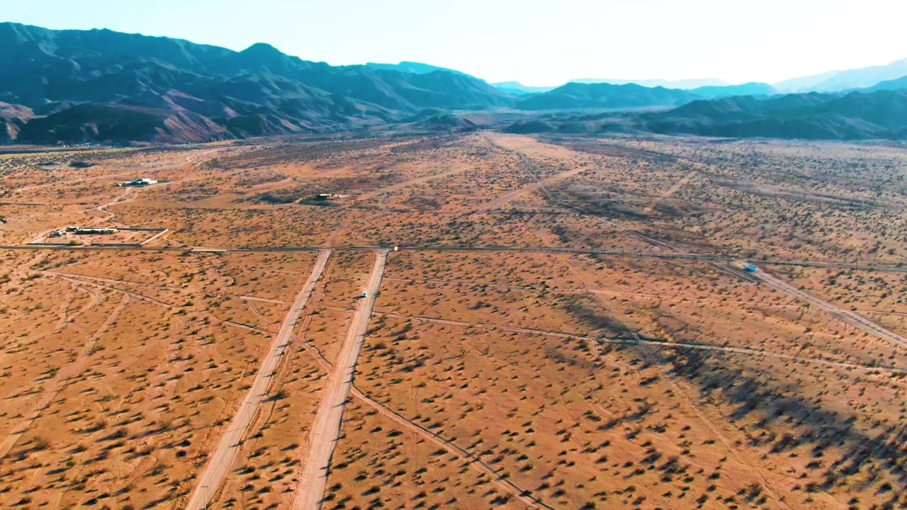 At 0:01, the aircraft is visible on the runway below the aerial camera, with parallel dirt roads, scattered brush, low buildings, mountain ridges, and no visible people.