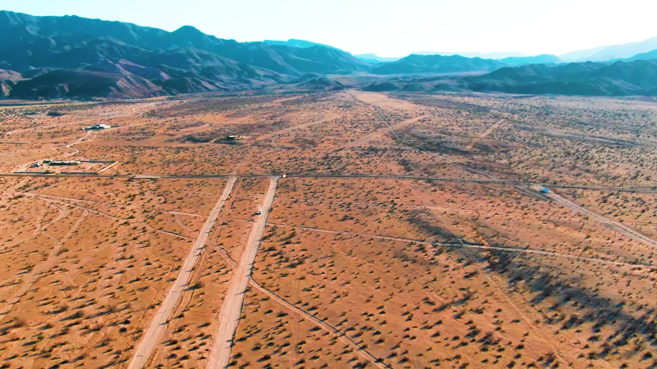 At 0:02, the runway stretches from the lower frame toward a small aircraft in the midground, surrounded by orange desert terrain, faint tracks, mountains, and no visible people.