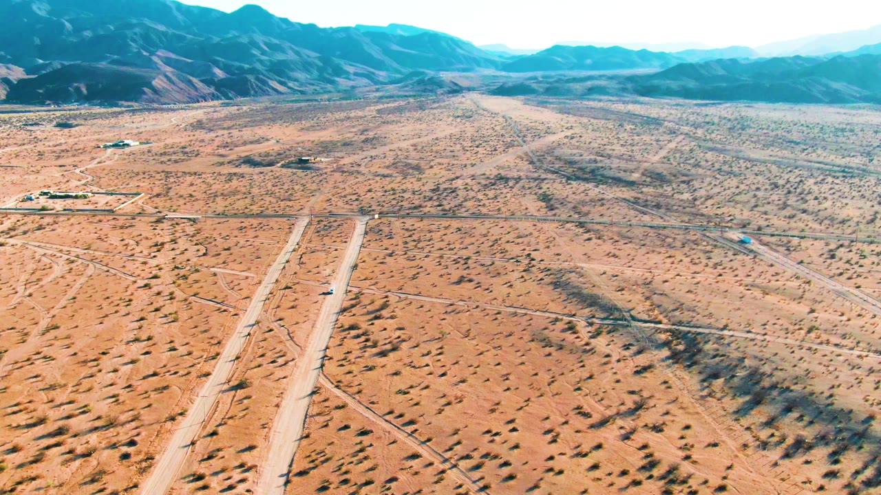 At 0:00, an aerial view over Bahia De Los Angeles shows a pale desert runway, a small car near the lower center, scattered scrub, distant buildings, blue-green mountains, and no visible people.