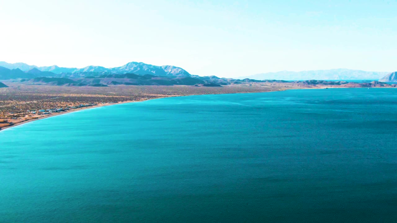 Aerial frame over Bahia De Los Angeles showing a curving shoreline at left, turquoise water across the foreground, islands, distant mountains, and no visible people.