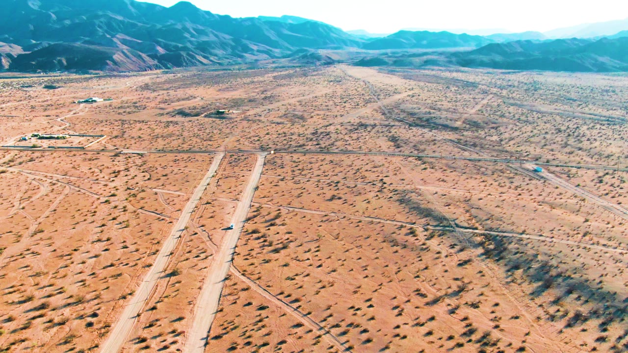 Aerial view over Bahia De Los Angeles, Mexico with the vehicle slightly farther along the pale runway, scrub-covered desert in the foreground, low buildings in the distance, mountains, and no visible people.