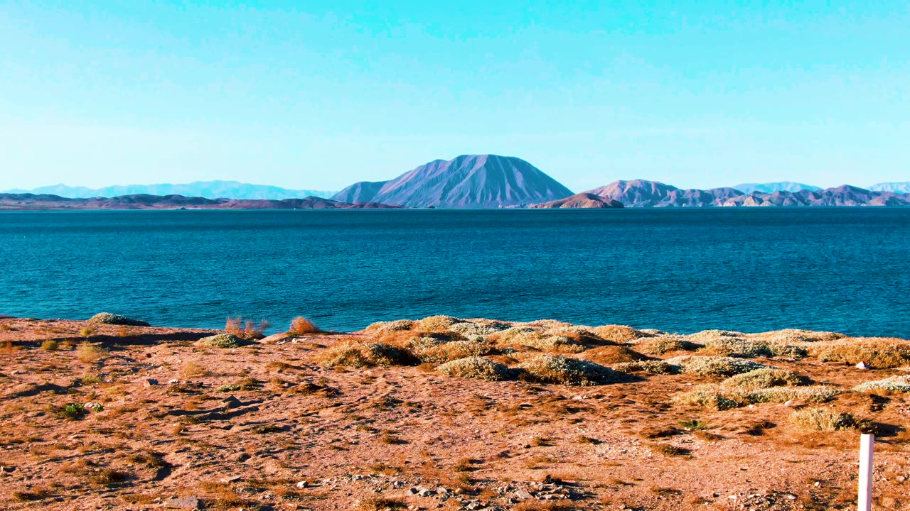 Wide view of Bahia De Los Angeles with dry rocky foreground, calm blue water, distant mountains, clear sky, and no visible people at the start of the clip.