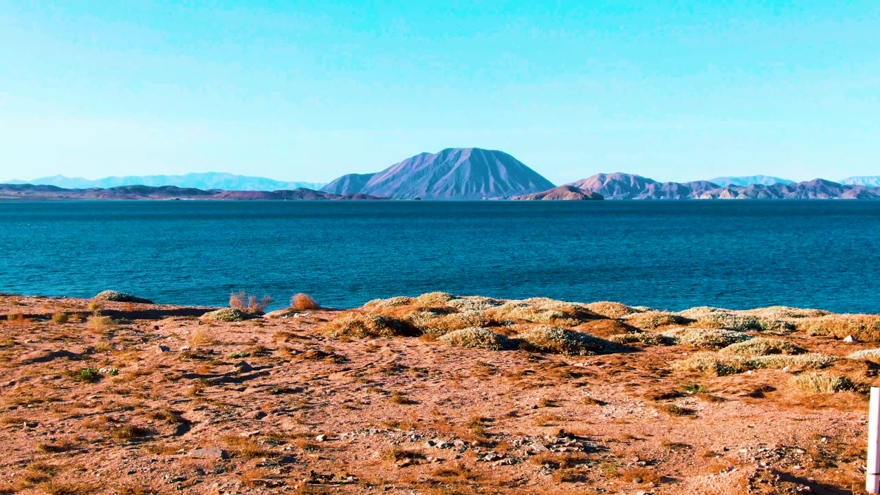 Wide Bahia De Los Angeles landscape with ochre rocky shore, blue water, a rounded distant mountain near center, pale ridges, clear sky, and no visible people.