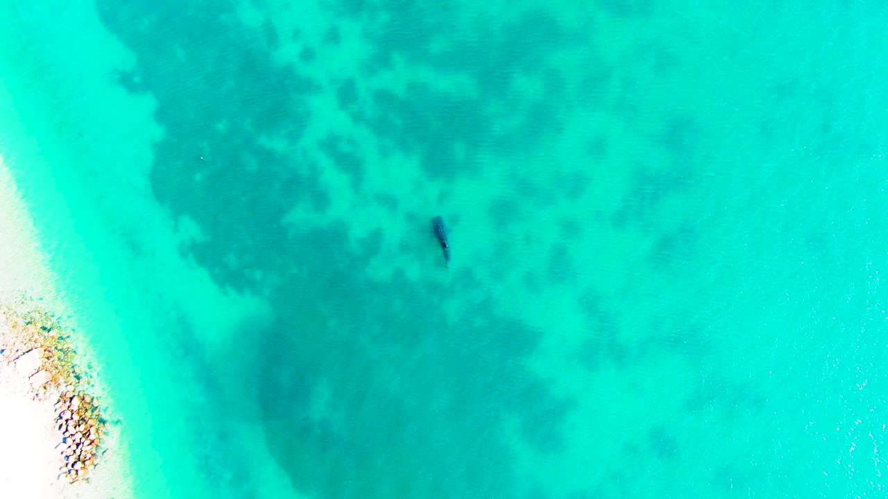Aerial frame over Bahia De Los Angeles shallows shows a narrow sandy edge with rocks at left, mottled darker seabed through turquoise water, and no recognizable people visible.