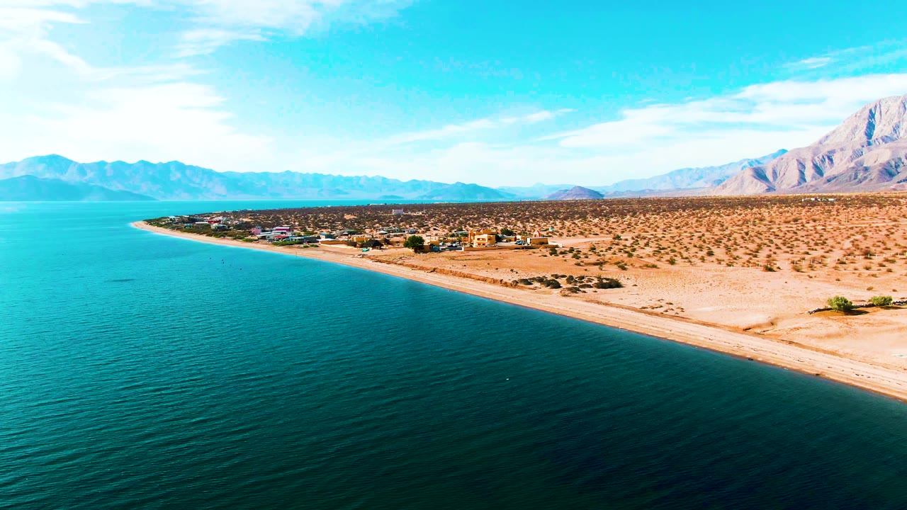 Aerial frame over Bahia De Los Angeles showing blue water in the foreground, a narrow sandy shoreline, arid terrain, distant mountains, and no visible people.
