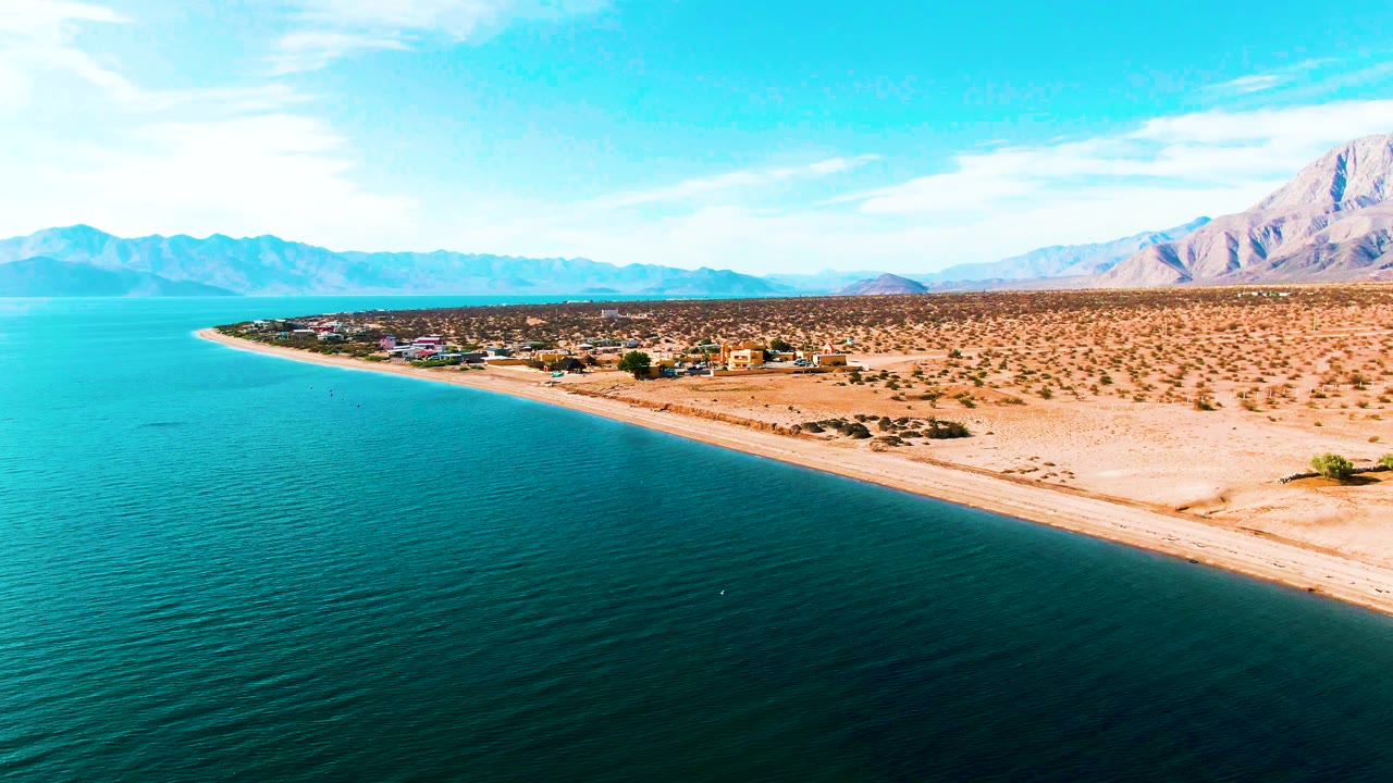 Blue coastal water borders a sandy beach with desert scrub and mountains beyond.
