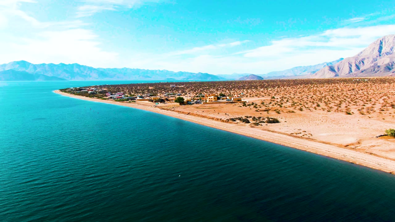 Aerial view of Bahia De Los Angeles with turquoise shallows near the beach, darker water in the foreground, sandy desert land, a small shoreline settlement, mountain backdrop, and no visible people.