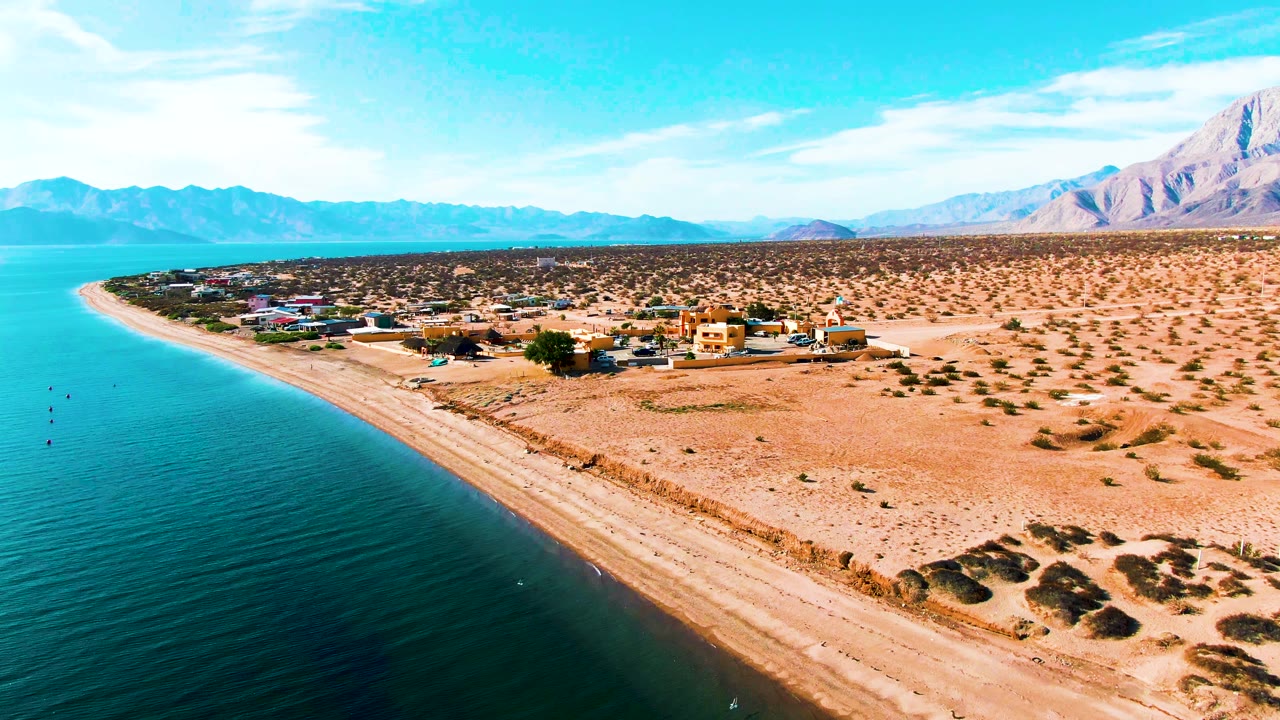 Aerial view over Bahia De Los Angeles with turquoise water at left, a sandy beach curving past low buildings, desert scrub, distant mountains, and no visible people.
