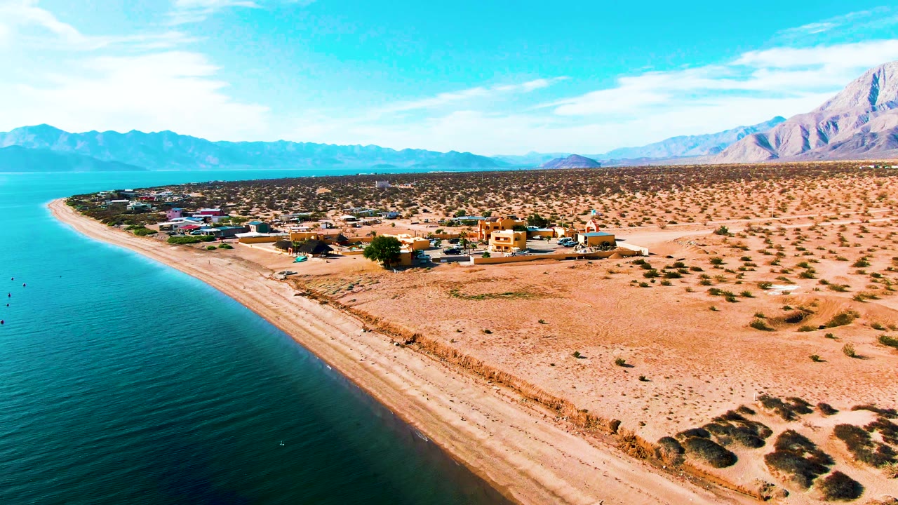 Wide drone-style frame of Bahia De Los Angeles showing blue water along the beach, scattered low buildings on arid ground, mountain ridges, and no visible people.
