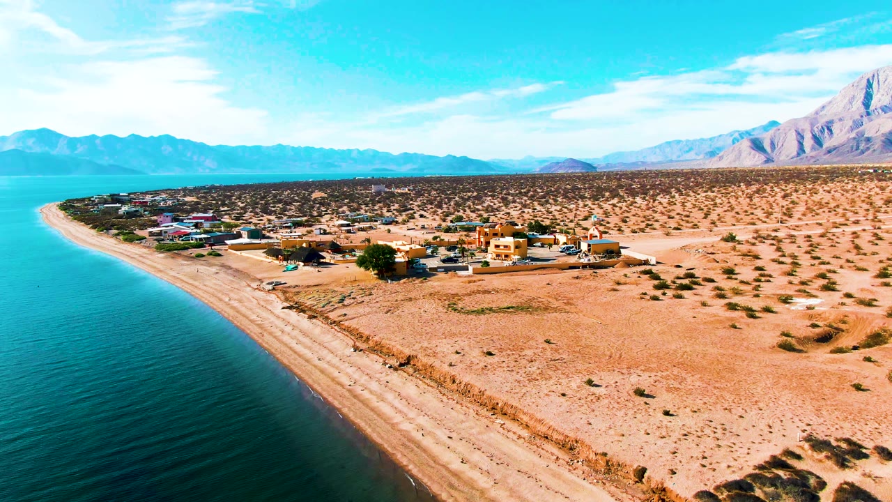 Aerial view in Bahia De Los Angeles with the bay filling the left side, a pale sandy shoreline, desert lots with scattered buildings, mountains on the horizon, and no visible people.