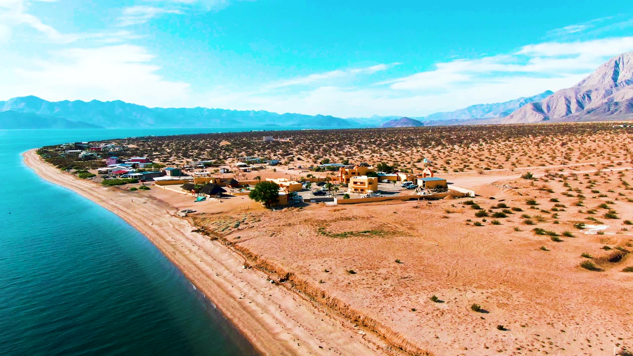 Curving beach beside calm water with desert buildings and mountains in the background.