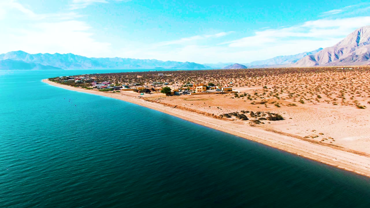 Opening aerial frame over Bahia De Los Angeles, Mexico, showing turquoise water, a sandy desert beach, low buildings inland, distant mountains, and no visible people.