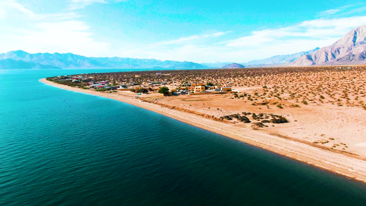 Drone view along a curving sandy shore with blue water and desert mountains.