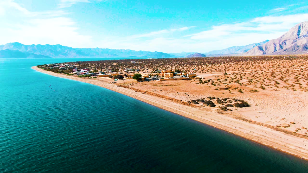 Aerial frame in Bahia De Los Angeles, Mexico, showing darker teal foreground water, a pale beach line, scattered desert vegetation, coastal buildings, mountains, and no visible people.