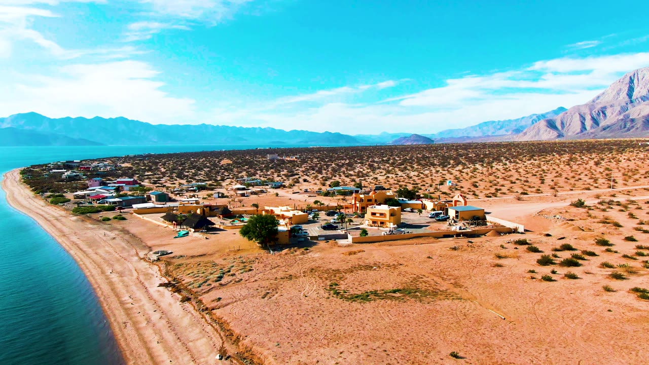 Aerial frame over Bahia De Los Angeles showing blue water and sandy beach at left, low village buildings near the shore, desert scrub inland, distant mountains, and no visible people.