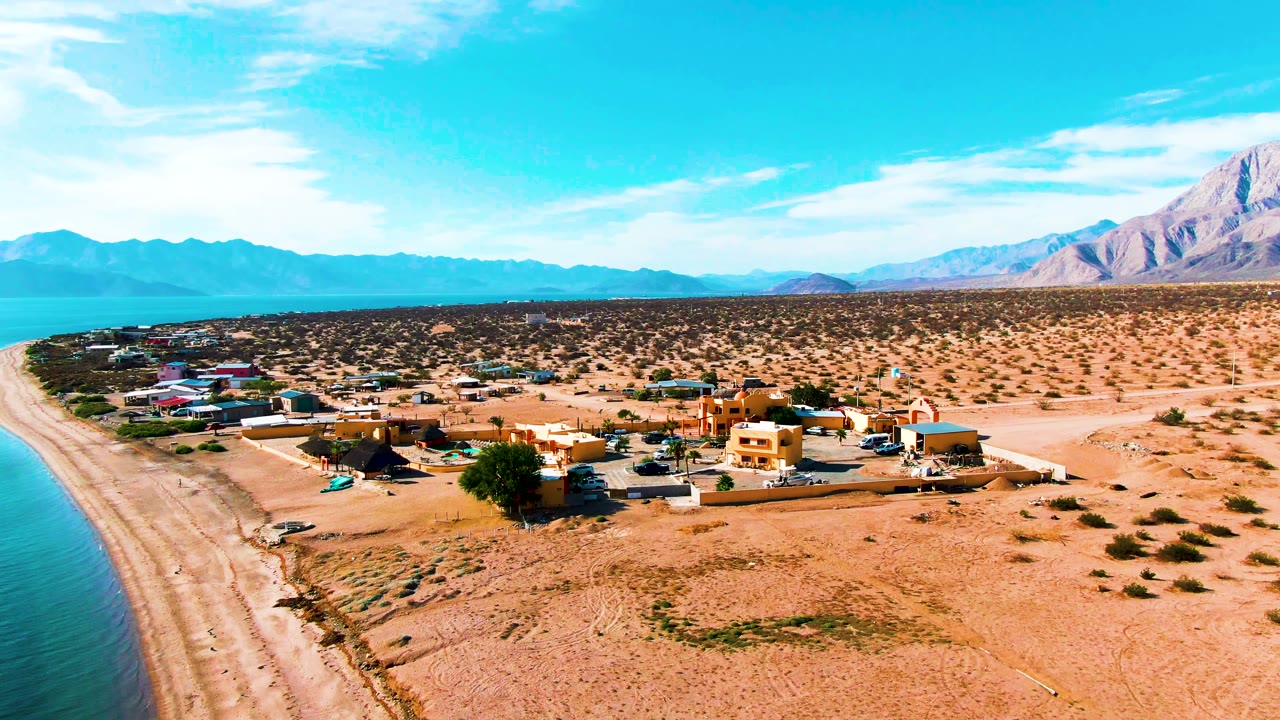Drone view of shoreline homes, sandy desert lots, blue water, and mountain horizon.